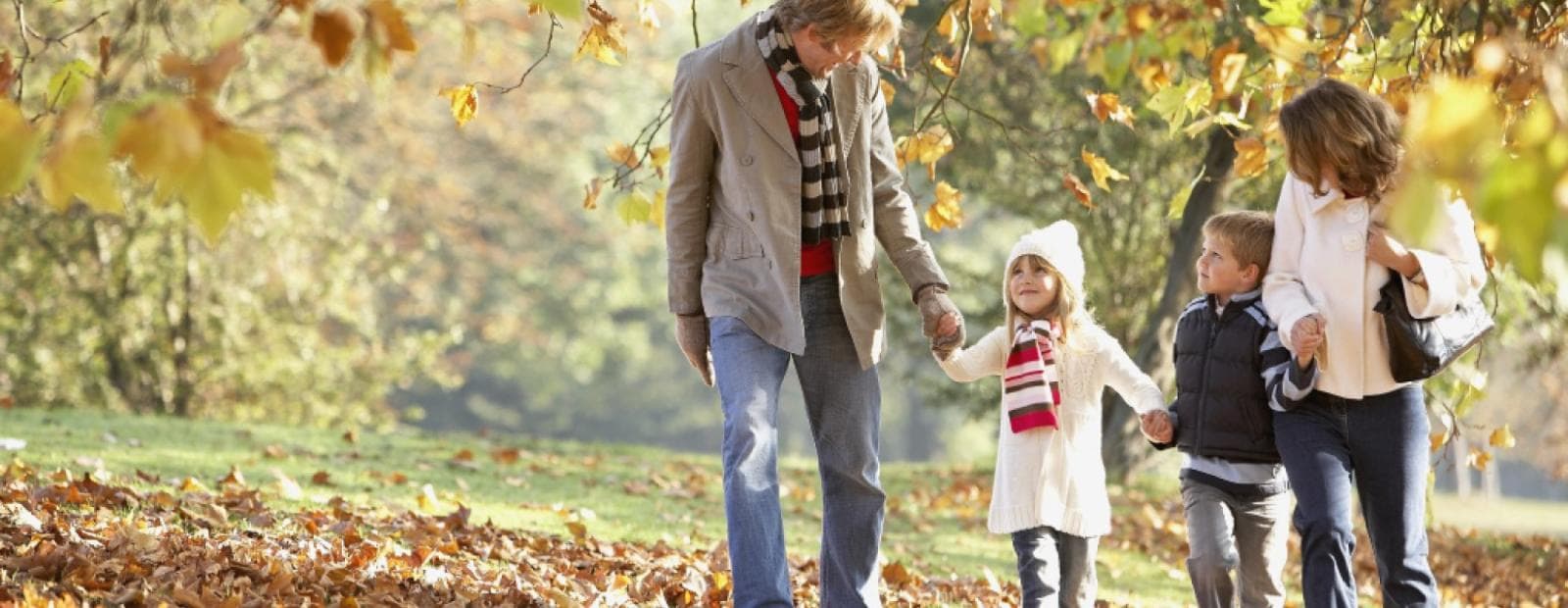 Family of four walking on a leafy path in autumn, surrounded by trees with colorful foliage.