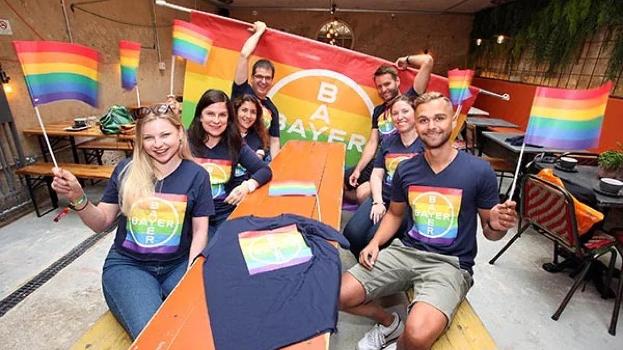 A group of people sitting at a table, holding rainbow flags and a large rainbow Bayer flag, wearing matching rainbow Bayer logo shirts.