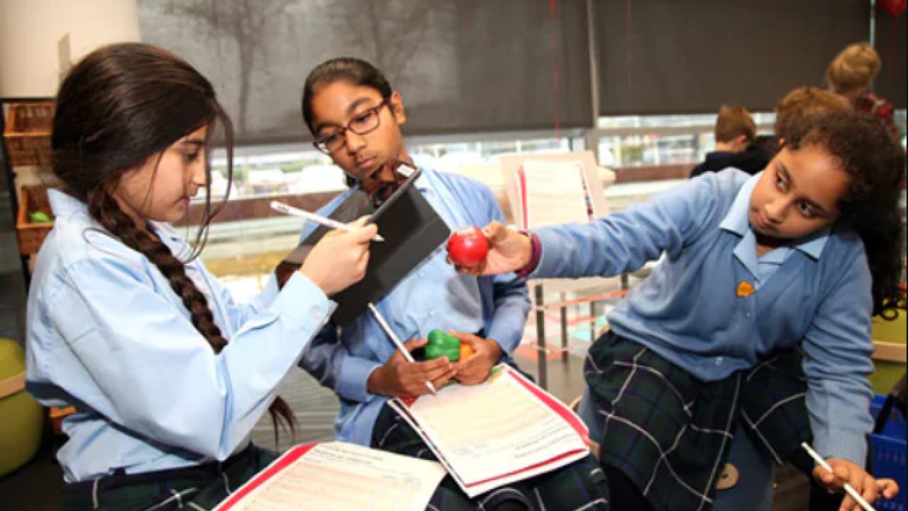 Three students in blue uniforms work together; one holds an apple, another writes on a clipboard, and the third observes. They stand in a classroom setting.