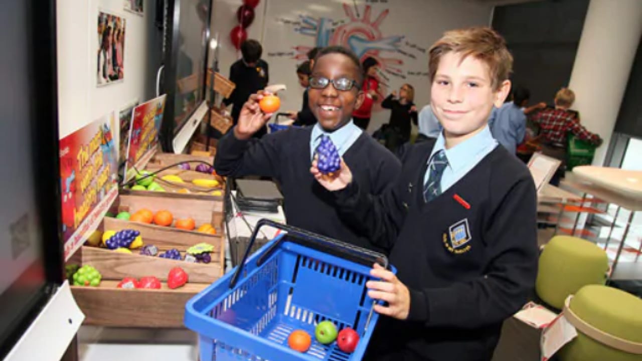 Two boys in school uniforms smile while holding colorful toy fruits and vegetables at a market stall display. One boy holds a shopping basket.