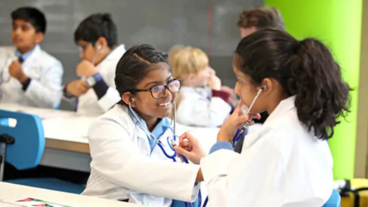 Children in white coats use stethoscopes in a classroom, engaging in a hands-on learning activity.