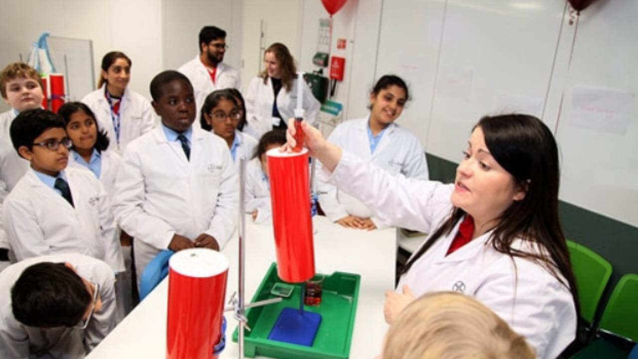 A group of students in lab coats gather around a woman demonstrating an experiment with a red cylinder in a science classroom.