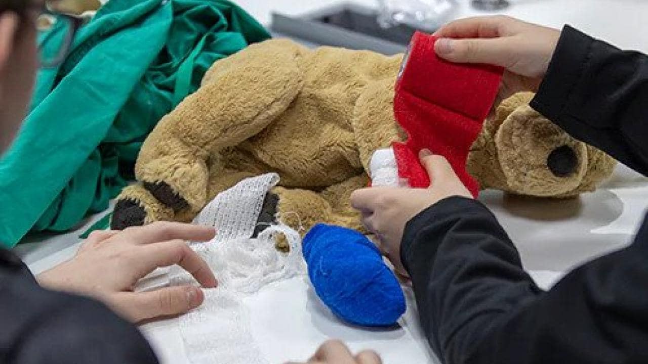 Hands are seen wrapping a plush teddy bear with red and white bandages on a table, surrounded by other medical supplies and green cloth.