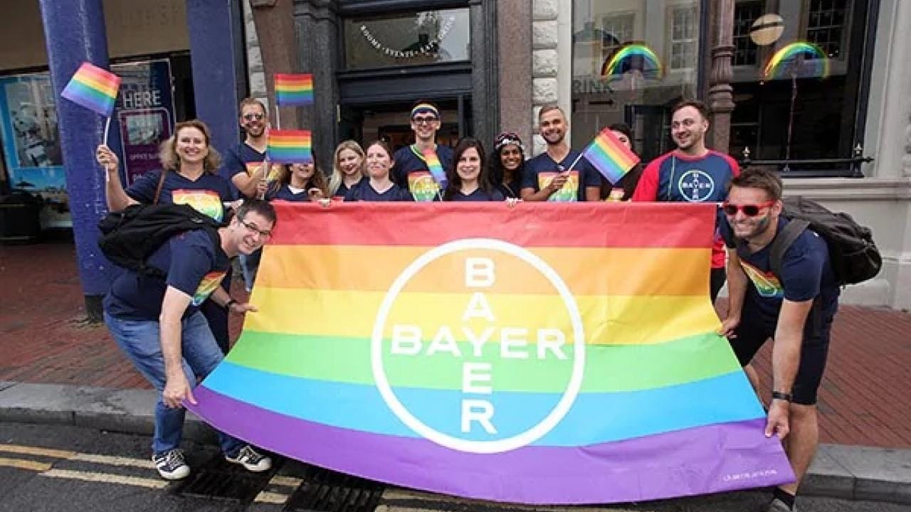 A group of people smiling and holding a large rainbow flag with "Bayer" written on it, standing on a city street. Some are wearing rainbow accessories.