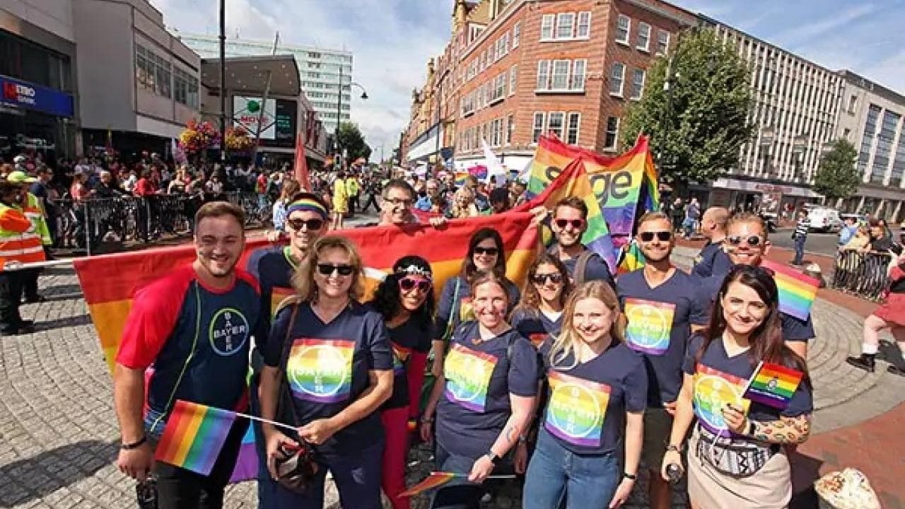 A diverse group of people wearing rainbow-themed shirts pose together with pride flags on a city street during a parade.