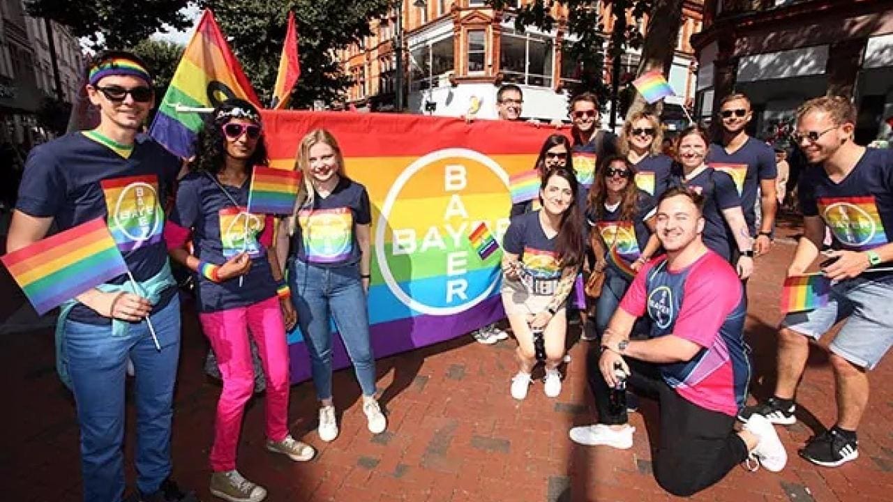 A group of people hold rainbow flags and a Bayer banner with a rainbow design during a street event.