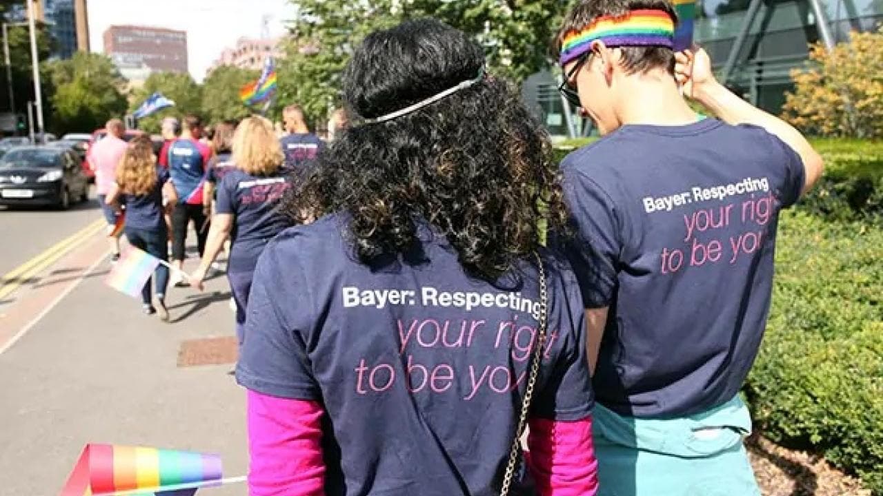 People walking in a parade wearing T-shirts that say "Bayer: Respecting your right to be you," with rainbow flags and accessories.