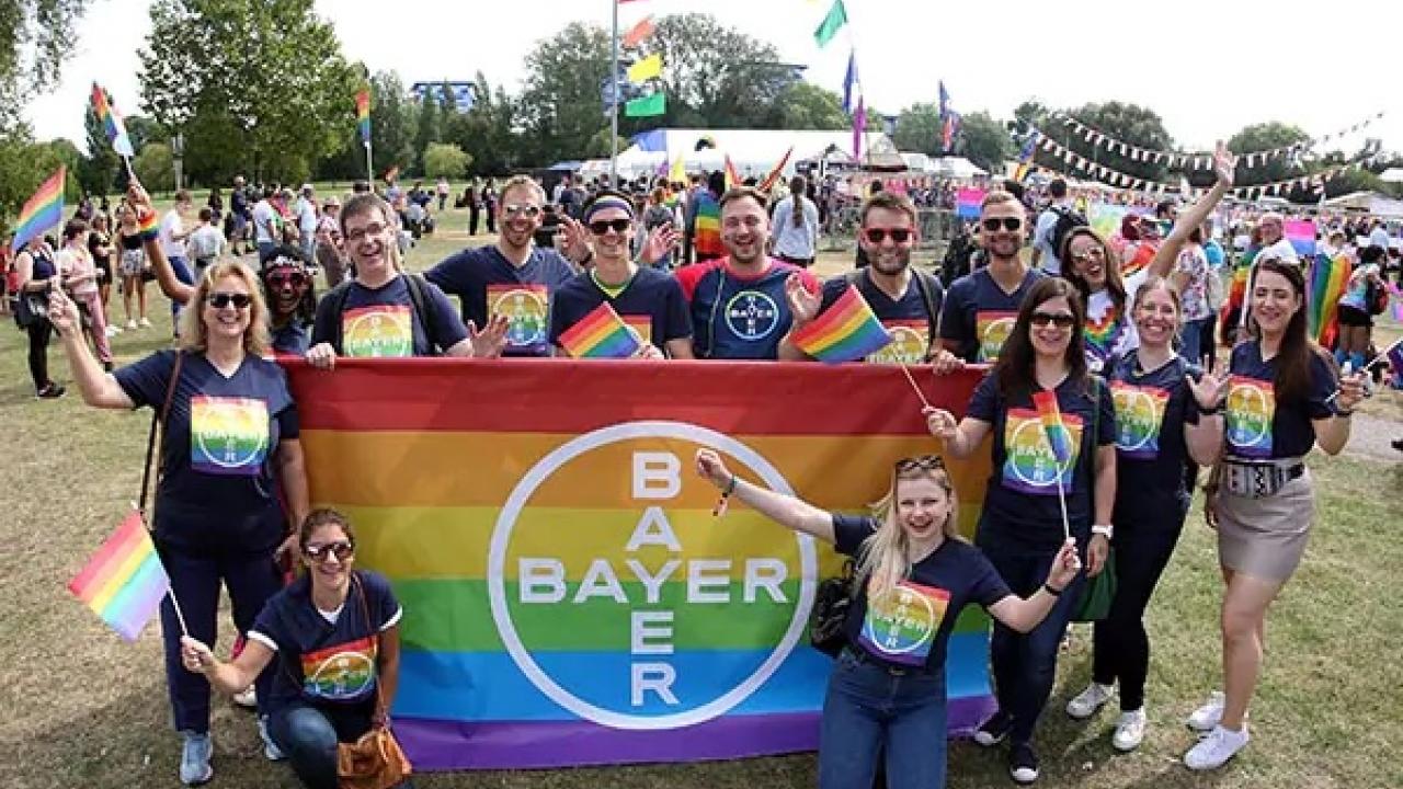 A group of people holding a Bayer banner with rainbow colors at an outdoor event. They are wearing matching rainbow-themed shirts and holding pride flags.