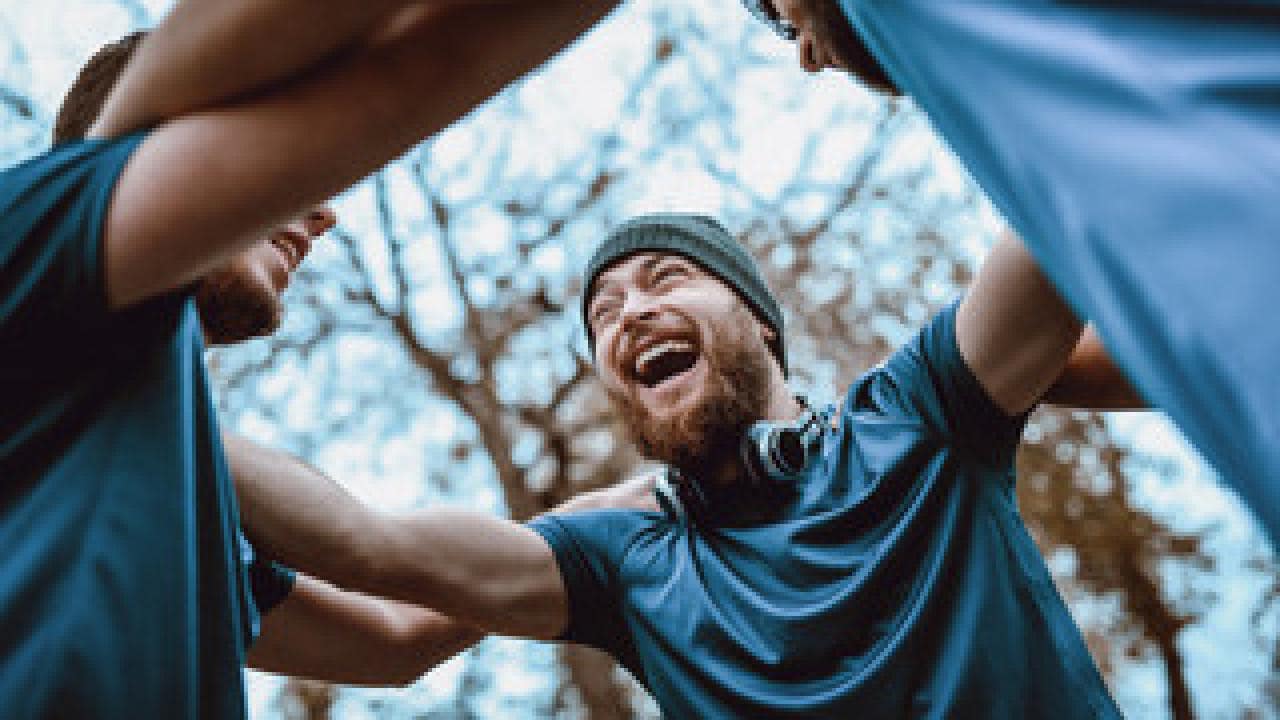 A group of people wearing blue shirts and a beanie stand in a circle outdoors, smiling and putting their arms around each other's shoulders.