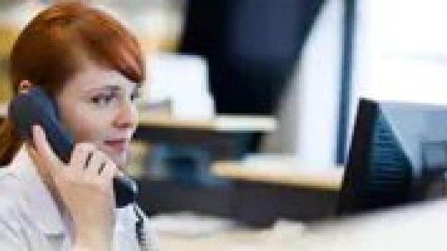 A person with red hair is speaking on a phone while looking at a computer screen in an office setting.