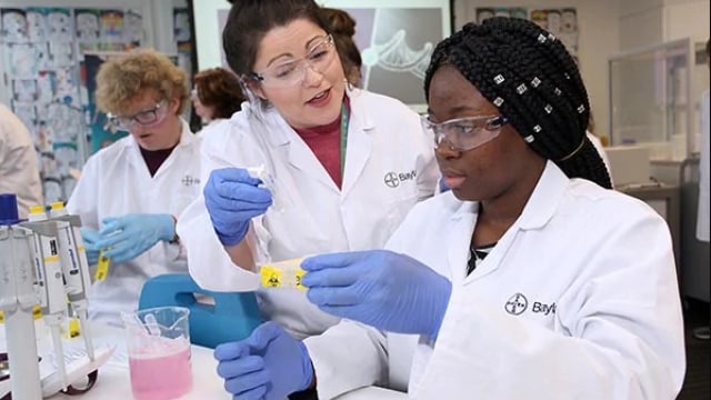 Two scientists in lab coats and goggles work with test samples. One is guiding the other, who is holding a vial. Additional scientists are working in the background.