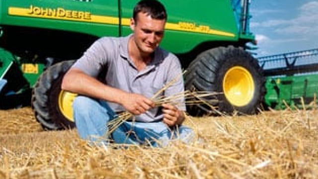 A person sits in a field of straw, examining some stalks, with a green John Deere combine harvester in the background.