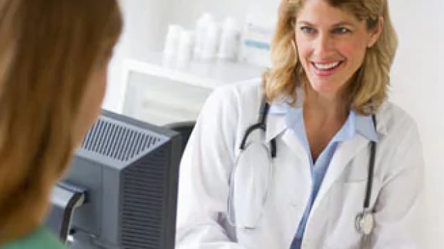 A doctor in a white coat with a stethoscope smiles while talking to a patient.