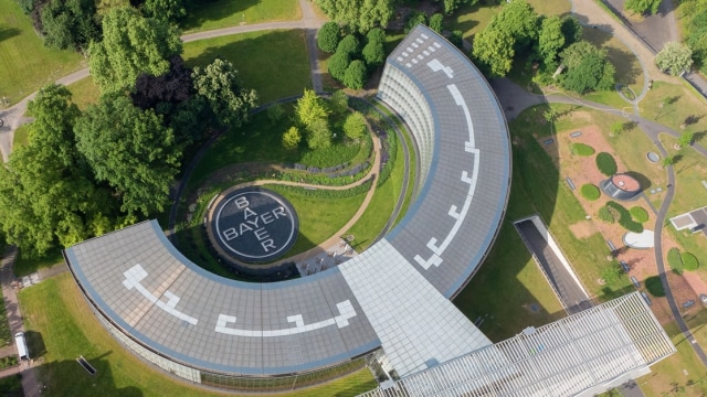 Aerial view of a U-shaped building with a circular garden in the center, featuring the Bayer logo. Surrounded by trees and a pathway.