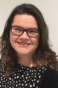 A woman with brown hair and glasses smiles while wearing a black polka dot top.