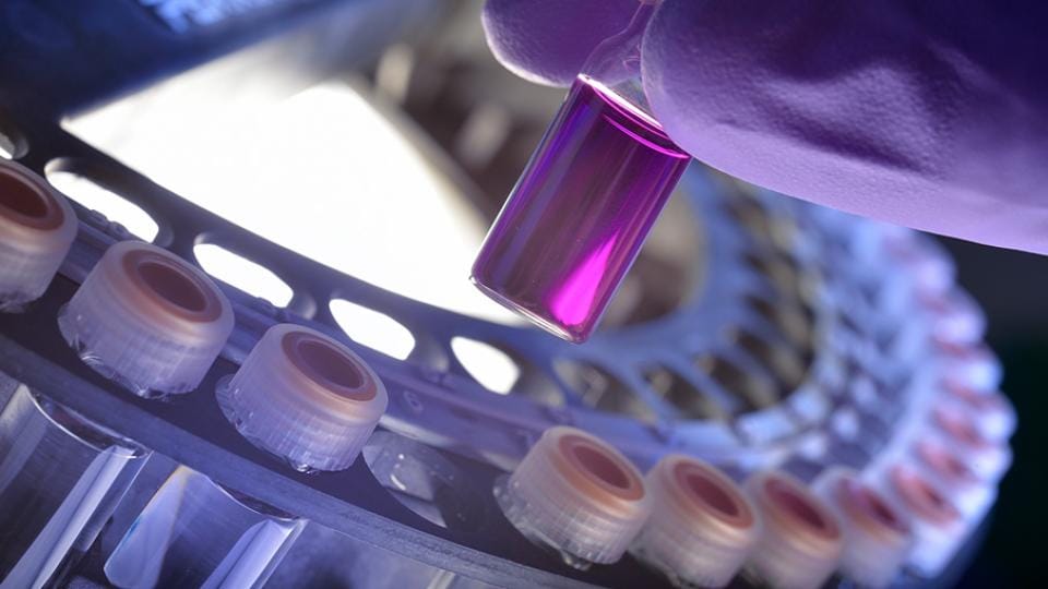A gloved hand holds a small vial of purple liquid near a lab centrifuge with several capped vials.